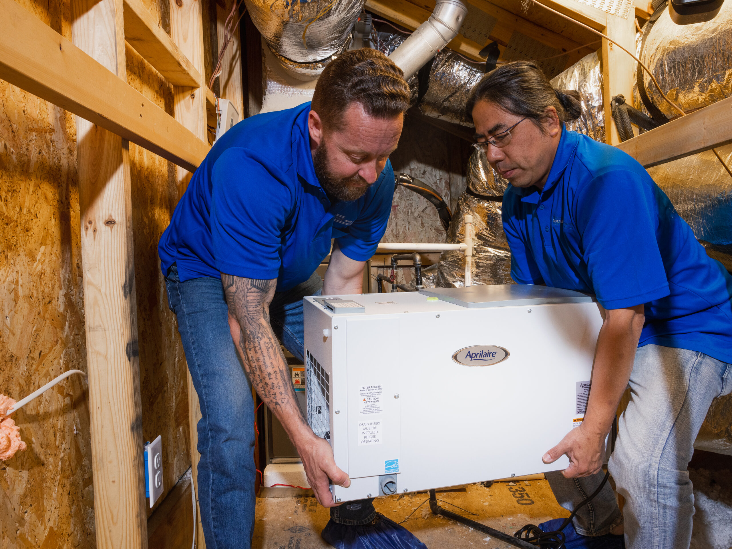 Two HVAC technicians in blue shirts install an Aprilaire unit in a residential space.